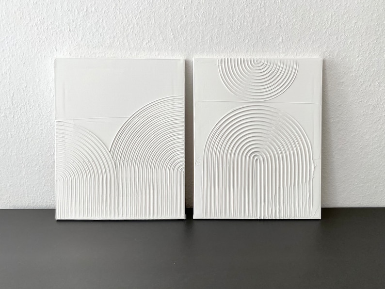 May include: Two white square canvases with raised, textured, white geometric shapes. The shapes are semi-circles and arcs, creating a modern abstract design.