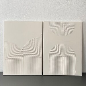 May include: Two white canvas panels with raised, textured, geometric designs. The designs are made of concentric circles and arcs, creating a modern and minimalist aesthetic.