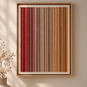 Pode incluir: Uma peça de arte de parede retangular com fios verticais em um gradiente de cores que variam do vermelho profundo e bordô à esquerda até tons de marrom, bege e amarelo brilhante à direita, emoldurada em uma moldura de madeira clara.