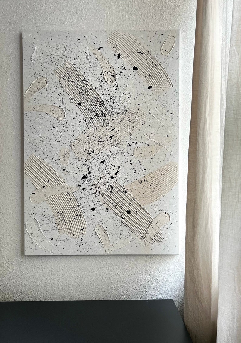 以下が含まれることがあります： 白、黒、ベージュの塗料を使用したキャンバス上の抽象画。絵画はテクスチャのある表面を持ち、さまざまな筆跡と飛沫を特徴としています。