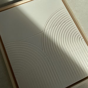 Pode incluir: Uma peça de arte de parede abstrata enquadrada com um design em relevo de círculos concêntricos sobrepostos em uma cor bege claro.