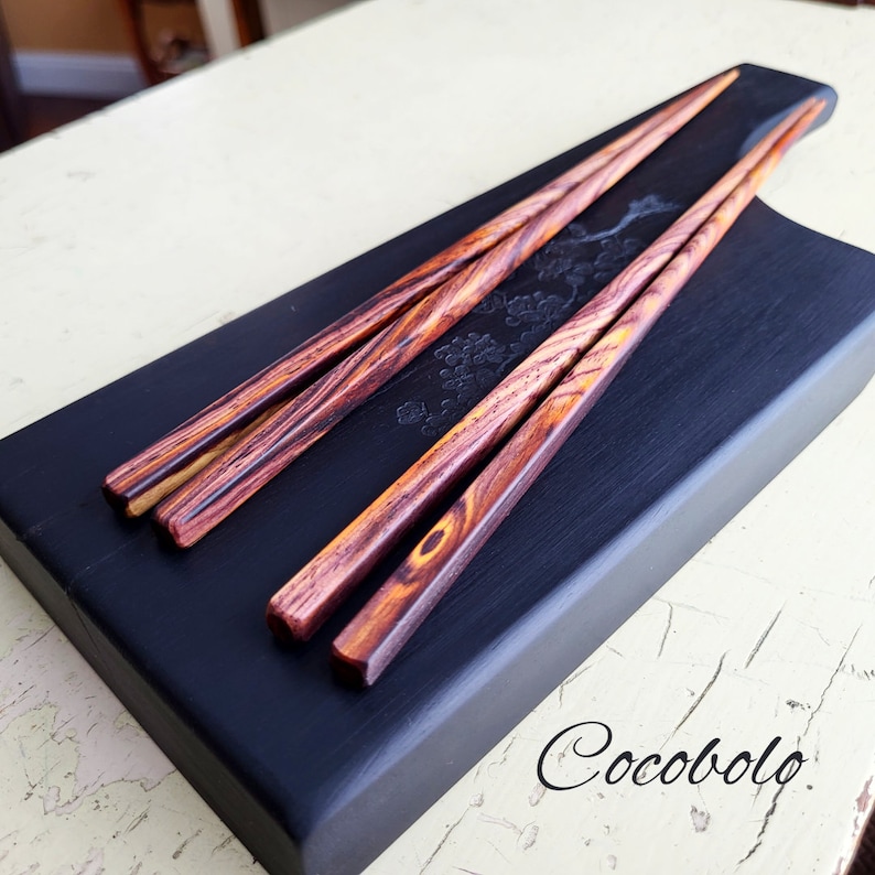 May include: Three dark brown wooden chopsticks with a reddish-brown hue and a natural wood grain pattern, resting on a black wooden serving tray with a carved floral design. The tray is labeled "Cocobolo".
