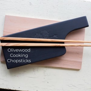 May include: Olivewood cooking chopsticks on a black serving board with a tree design. The board is on a light wood surface.