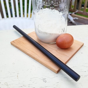 May include: A black rolling pin, a brown egg, and a glass jar filled with white flour on a wooden chopping board.