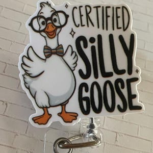 May include: A white badge reel featuring a cartoon goose wearing glasses and a bow tie. The text "CERTIFIED SILLY GOOSE" is printed in black. The badge reel has a clear plastic base and a metal clip.