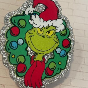 May include: A decorative ornament featuring a green character wearing a red Santa hat and a red scarf, positioned in the center of a green Christmas wreath adorned with red and blue ornaments, all set against a silver glitter border background.