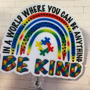 May include: A white badge holder with a rainbow and puzzle pieces. The text "In a world where you can be anything, be kind" is written around the rainbow. The rainbow is made of red, orange, yellow, green, blue, and purple. The puzzle pieces are blue, green, yellow, and red.