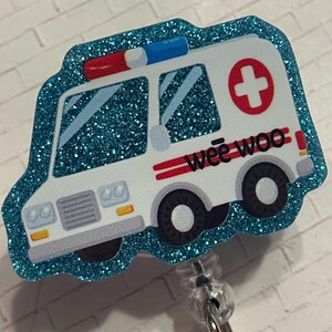 May include: A badge reel featuring a white ambulance with a blue glitter border, red and blue emergency lights, a red cross symbol, and the text "wee woo" in black lettering. The ambulance has black wheels and yellow headlights on the front bumper.