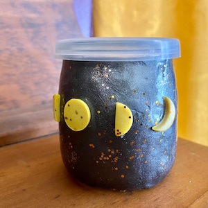 Puede incluir: Un tarro de vidrio negro con una tapa transparente decorada con purpurina dorada y fases lunares amarillas. El tarro está sobre una superficie de madera.
