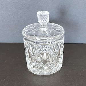 Może przedstawiać: Przezroczysty, szlifowany szklany słoik z pokrywką. Słoik ma geometryczny wzór i mały, diamentowy uchwyt. Cylindryczny słoik stoi na ciemnej powierzchni. Pokrywka dobrze pasuje.