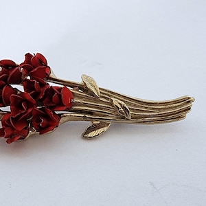 May include: A gold-toned brooch with a cluster of red enamel flowers. The flowers are arranged in a bouquet shape with stems and leaves.