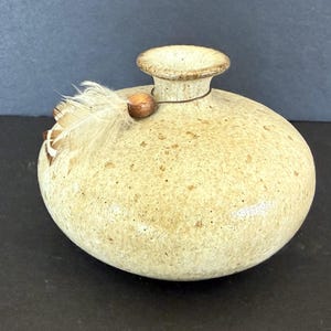 May include: A round, beige ceramic vase with a speckled finish and a narrow neck. A thin cord is tied around the neck, featuring a single brown wooden bead and a small, light-colored feather attached to the side of the vase's rounded body.