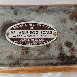 Peut inclure: Une balance &agrave; &oelig;ufs en m&eacute;tal vintage avec une &eacute;tiquette ovale brune qui indique "Reliable Mfg. Division of Reliable Egg Scale - See Weight Chart - James Mfg. Co. Los Angeles, Calif. U.S.A."