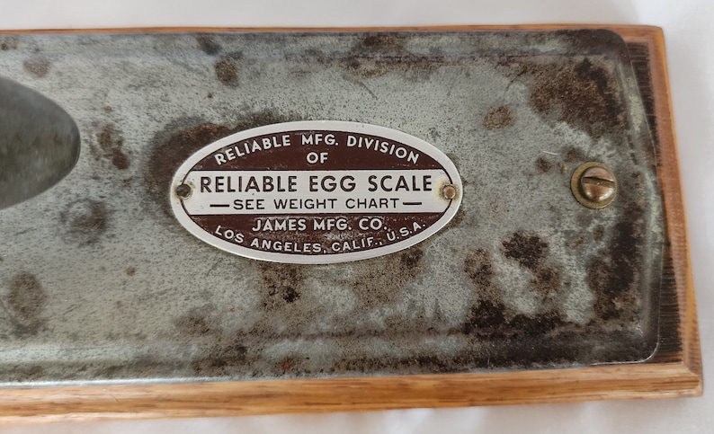 Peut inclure: Une balance &agrave; &oelig;ufs en m&eacute;tal vintage avec une &eacute;tiquette ovale brune qui indique "Reliable Mfg. Division of Reliable Egg Scale - See Weight Chart - James Mfg. Co. Los Angeles, Calif. U.S.A."