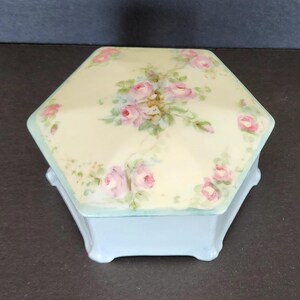 May include: A hexagonal porcelain trinket box with a light blue base and a cream lid. The lid is decorated with a hand-painted floral design of pink roses and green leaves.