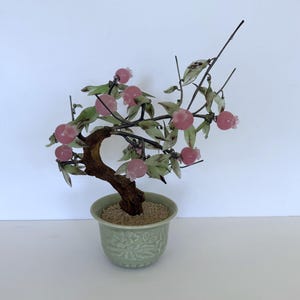 May include: A decorative glass bonsai tree with pink fruit and green leaves. The tree is in a light green ceramic pot with a textured design. The trunk is brown and the branches are dark.