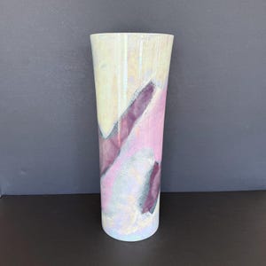 May include: A tall cylindrical vase featuring an iridescent finish with abstract color blocks in shades of pale yellow, soft pink, and deep purple. The surface has a smooth, glossy texture with subtle silver-toned speckled detailing throughout.