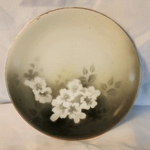 May include: A white porcelain plate with a black and white floral design. The flowers are white with black centers and are surrounded by black leaves. The plate has a gold rim.