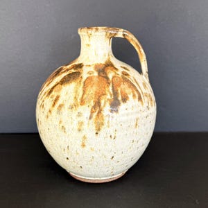 May include: A handmade ceramic pitcher with a rounded body and a handle. The pitcher is primarily off-white with brown and orange drip glaze patterns. The base is a reddish-brown color. The pitcher has a small spout.