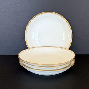 Puede incluir: Un juego de cuatro cuencos de porcelana blanca con borde dorado. Los cuencos están apilados y dispuestos sobre una superficie oscura. Los cuencos tienen un diseño simple y elegante.