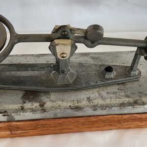 Peut inclure: Une balance &agrave; &oelig;ufs vintage en argent avec une base en bois. La balance poss&egrave;de un anneau m&eacute;tallique pour maintenir l'&oelig;uf et un levier pour le peser. La balance est &eacute;tiquet&eacute;e "Reliable Mfg. Division of Reliable Egg Scale See Weight Chart James Mfg. Co. Los Angeles, Calif. USA."