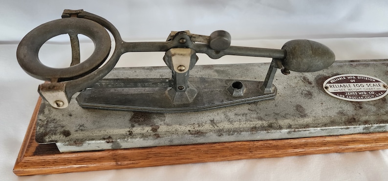 Peut inclure: Une balance &agrave; &oelig;ufs vintage en argent avec une base en bois. La balance poss&egrave;de un anneau m&eacute;tallique pour maintenir l'&oelig;uf et un levier pour le peser. La balance est &eacute;tiquet&eacute;e "Reliable Mfg. Division of Reliable Egg Scale See Weight Chart James Mfg. Co. Los Angeles, Calif. USA."