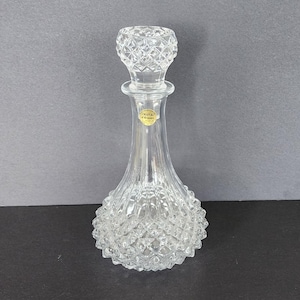 Könnte beinhalten: Eine klare Glaskaraffe mit einem strukturierten Diamantenmuster. Die Karaffe hat einen runden Stopfen mit einem ähnlichen Diamantenmuster. Das Etikett auf der Karaffe lautet "Cristal d'Arques".