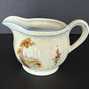 May include: Cream-colored ceramic creamer with a handle and spout, featuring a landscape scene with trees and a building. The rim is accented with a blue stripe, and the creamer has a floral design on the side.
