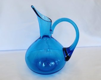 May include: A bright blue glass pitcher with a rounded body and a curved spout. The pitcher has a single handle.