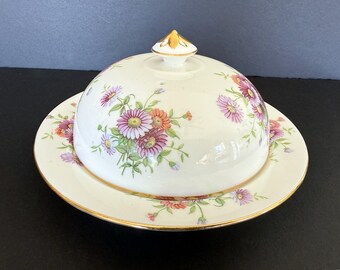 May include: A white ceramic butter dish with a domed lid and matching plate. The dish is decorated with a floral pattern in shades of pink, orange, and green, with gold trim. The lid has a gold finial.