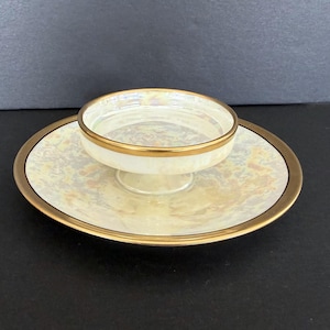 Puede incluir: Un juego de servir vintage de dos piezas con un cuenco pequeño con pedestal y un platillo a juego. Ambas piezas tienen un acabado cremoso e iridiscente con ribete dorado. El cuenco se asienta sobre el platillo, creando una presentación escalonada. El juego es probablemente para servir porciones pequeñas.