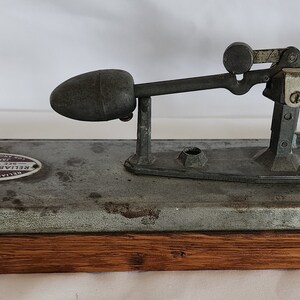 Peut inclure: Un testeur d'&oelig;ufs vintage en m&eacute;tal avec une base en bois. Le testeur a un bras en m&eacute;tal avec un porte-&oelig;uf lest&eacute; et une &eacute;chelle pour mesurer la fra&icirc;cheur de l'&oelig;uf.