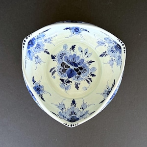 Puede incluir: Un cuenco de cerámica triangular de color crema con un diseño floral azul. El cuenco presenta una gran flor central y pequeños detalles florales alrededor del borde. Los bordes están festoneados con pequeños agujeros espaciados.