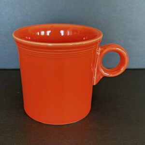 Könnte beinhalten: Eine leuchtend orangefarbene Keramik-Tasse mit einem abgerundeten Henkel. Die Tasse hat eine zylindrische Form mit einem leicht ausgestellten Rand und einer glatten, glänzenden Oberfläche. Die Tasse ist einfarbig leuchtend orange.