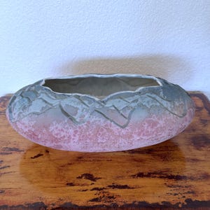 May include: An oval ceramic planter featuring a textured finish with a gradient of soft pink at the base transitioning into a muted sage green and grey at the top. The rim has a scalloped, uneven edge with a carved geometric mountain-like pattern.