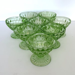Könnte beinhalten: Eine Sammlung von acht grünen Dessertschalen aus Glas. Jede Schale hat ein Diamantmuster auf der Schüssel und einen Sockel mit gerilltem Design. Die Schalen sind auf einer weißen Oberfläche angeordnet.