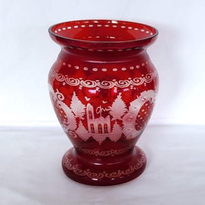 May include: A red glass vase with a white etched design of a house and trees. The vase has a round body and a flared lip.