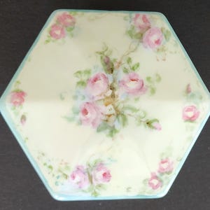 May include: A hexagonal porcelain trinket box with a light blue trim and a delicate floral design featuring pink roses and green leaves.