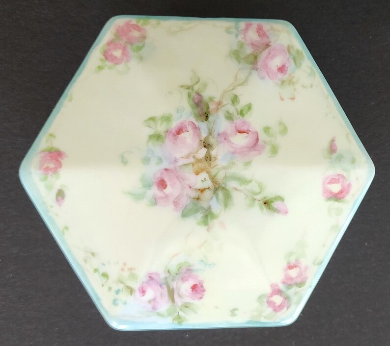 May include: A hexagonal porcelain trinket box with a light blue trim and a delicate floral design featuring pink roses and green leaves.