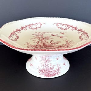 May include: White ceramic cake stand with a scalloped edge and a red toile pattern. The design features a pastoral scene with birds and trees. The base and rim are also decorated with the same red pattern.