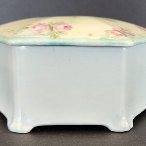 May include: A white porcelain octagonal trinket box with a floral design. The lid is decorated with pink roses and green leaves on a cream background. The box has a light blue trim around the top edge.