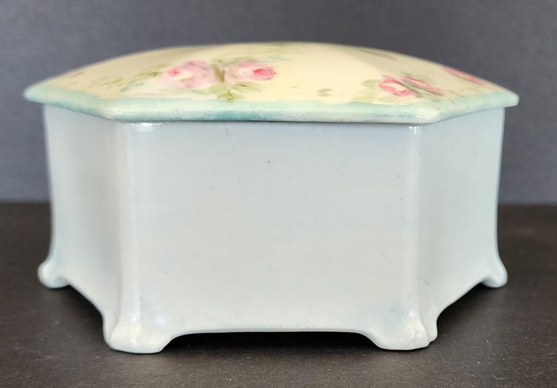 May include: A white porcelain octagonal trinket box with a floral design. The lid is decorated with pink roses and green leaves on a cream background. The box has a light blue trim around the top edge.