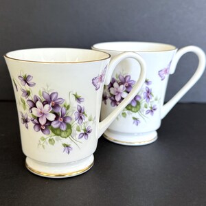 May include: Two white porcelain mugs featuring a delicate floral pattern of purple violets and green leaves, accented with small butterflies. Each mug has a flared rim and a base finished with a thin gold trim along the edges and handle.