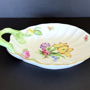 May include: A white porcelain serving dish with a scalloped edge and a green handle. The dish is decorated with a floral design featuring yellow, purple, pink, and blue flowers. A gold trim accents the edge.