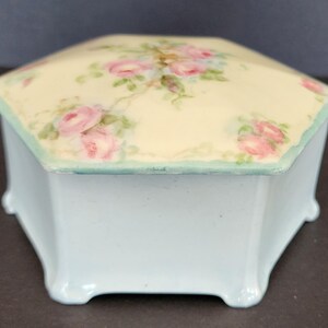 May include: A hexagonal porcelain trinket box with a light blue base and a white lid decorated with hand-painted pink roses and green leaves.