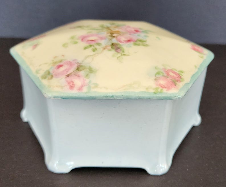 May include: A hexagonal porcelain trinket box with a light blue base and a white lid decorated with hand-painted pink roses and green leaves.