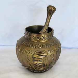 Puede incluir: Un mortero y un pilón de color dorado con un diseño en relieve y el nombre "Maimónides" inscrito en el lateral. El mortero también tiene los años "1135" y "1204" inscritos en él.