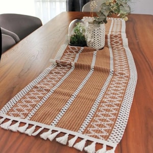 May include: A brown and white table runner with a striped and geometric design. The runner has white tassels at the ends and is placed on a wooden table. The runner is approximately 152 cm long.