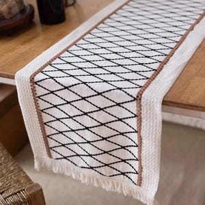 May include: A white table runner featuring a black diamond design and a brown border. The runner has a fringed edge and is placed on a wooden table. This decorative runner adds a touch of elegance to any dining setting.