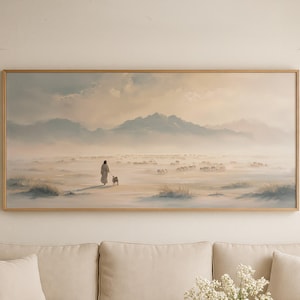 Könnte beinhalten: Ein gerahmtes Landschaftsgemälde zeigt eine Person mit einem Hund, die durch ein nebliges Feld auf entfernte Berge zugeht. Das Kunstwerk wird über einem beigen Sofa mit passenden Kissen, einem Holztisch und einer Blumenvase ausgestellt.